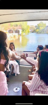 Group of friends toasting with champagne on a covered pontoon boat, cruising a sunny lake past waterfront cottages and green trees