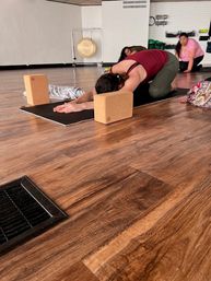 Person in child's pose using cork yoga blocks on a black mat in a bright yoga studio with wooden floor; two classmates and a gong in the background.