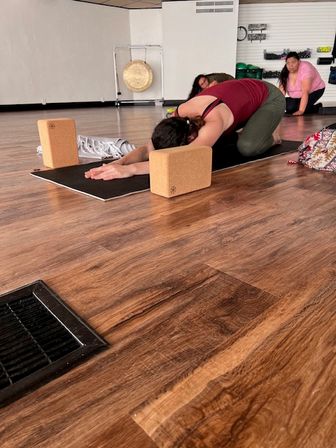 Person in child's pose using cork yoga blocks on a black mat in a bright yoga studio with wooden floor; two classmates and a gong in the background.