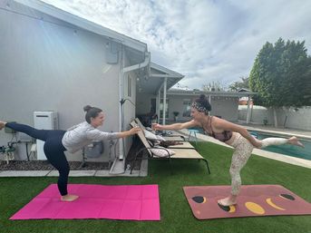 Two women balancing in standing bow yoga poses on pink and patterned mats, reaching forward to clink drinks in a sunny suburban backyard next to a pool.