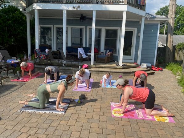 Outdoor yoga class on a sunny suburban backyard patio with several people on colorful mats practicing tabletop/cat-cow poses in front of a two-story house