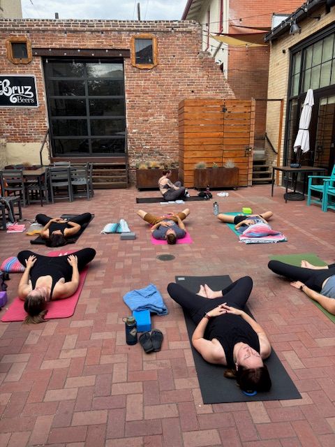 Group outdoor yoga class on colorful mats in an urban brick courtyard, participants lying on their backs in relaxation pose near patio furniture and wooden accents