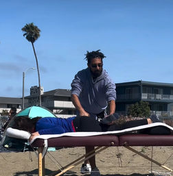 Seaside beach massage on a portable table: therapist stretching a client’s leg on a sandy shore with palm tree and coastal condos under a clear blue sky