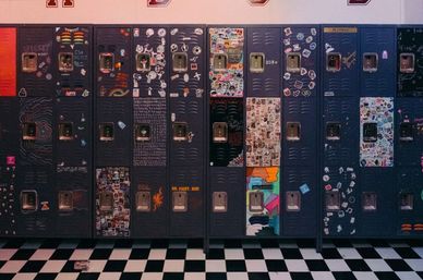 Row of dark metal school lockers covered in colorful stickers, doodles and collages in a lively high school hallway with a black-and-white checkerboard floor and a discarded soda can