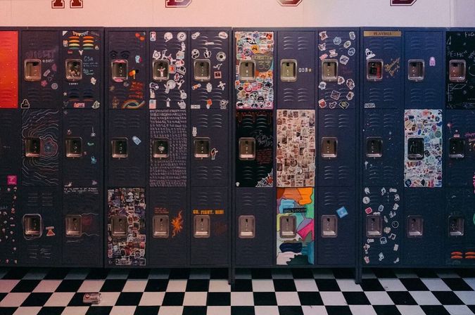Row of dark metal school lockers covered in colorful stickers, doodles and collages in a lively high school hallway with a black-and-white checkerboard floor and a discarded soda can