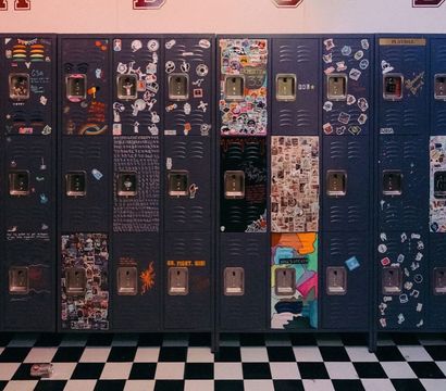 Row of dark metal school lockers covered in colorful stickers, doodles and collages in a lively high school hallway with a black-and-white checkerboard floor and a discarded soda can