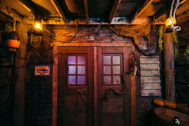 Dimly lit rustic boathouse interior with weathered double wooden doors and frosted panes, warm lantern light, hanging fishing nets, ropes, buoys and maritime decor