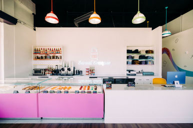Bright pastel dessert shop interior with a pink glass pastry case of donuts and pastries, marble service counter with register, shelves of syrups and merch, a neon wall sign and colorful pendant lights.