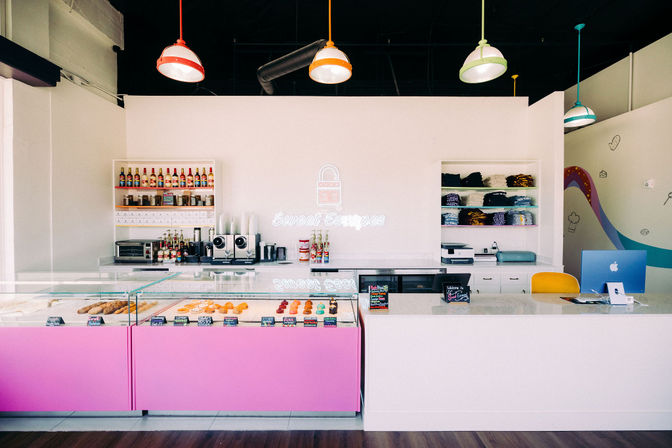 Bright pastel dessert shop interior with a pink glass pastry case of donuts and pastries, marble service counter with register, shelves of syrups and merch, a neon wall sign and colorful pendant lights.
