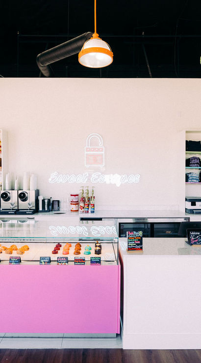 Bright pastel dessert shop interior with a pink glass pastry case of donuts and pastries, marble service counter with register, shelves of syrups and merch, a neon wall sign and colorful pendant lights.