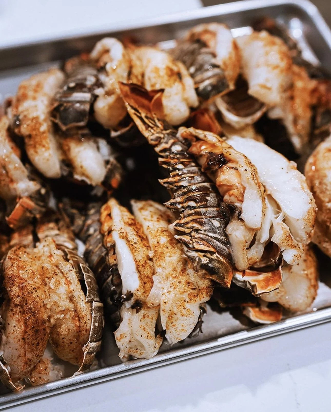 Close-up of seasoned grilled lobster tails on a metal tray — juicy white meat and charred shells ready for a seafood feast.