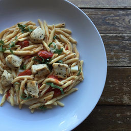 Top-down view of a white bowl of twisted pasta with grilled chicken cubes, roasted cherry tomatoes and fresh basil on a rustic wooden table