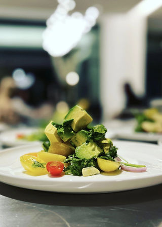 Close-up of fresh avocado and baby potato salad — stacked avocado cubes, cherry tomatoes, mixed greens and red onion on a white plate in a modern restaurant setting