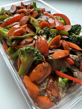 Colorful Asian-style beef stir-fry with tender beef, sliced carrots, broccoli florets and red and green bell peppers served over white rice in a clear takeout container