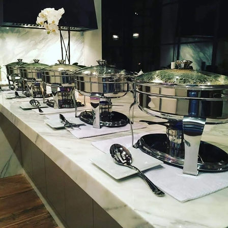 Sleek stainless-steel chafing dishes lined on a marble buffet with serving spoons and a white orchid accent — elegant hotel or event catering setup.