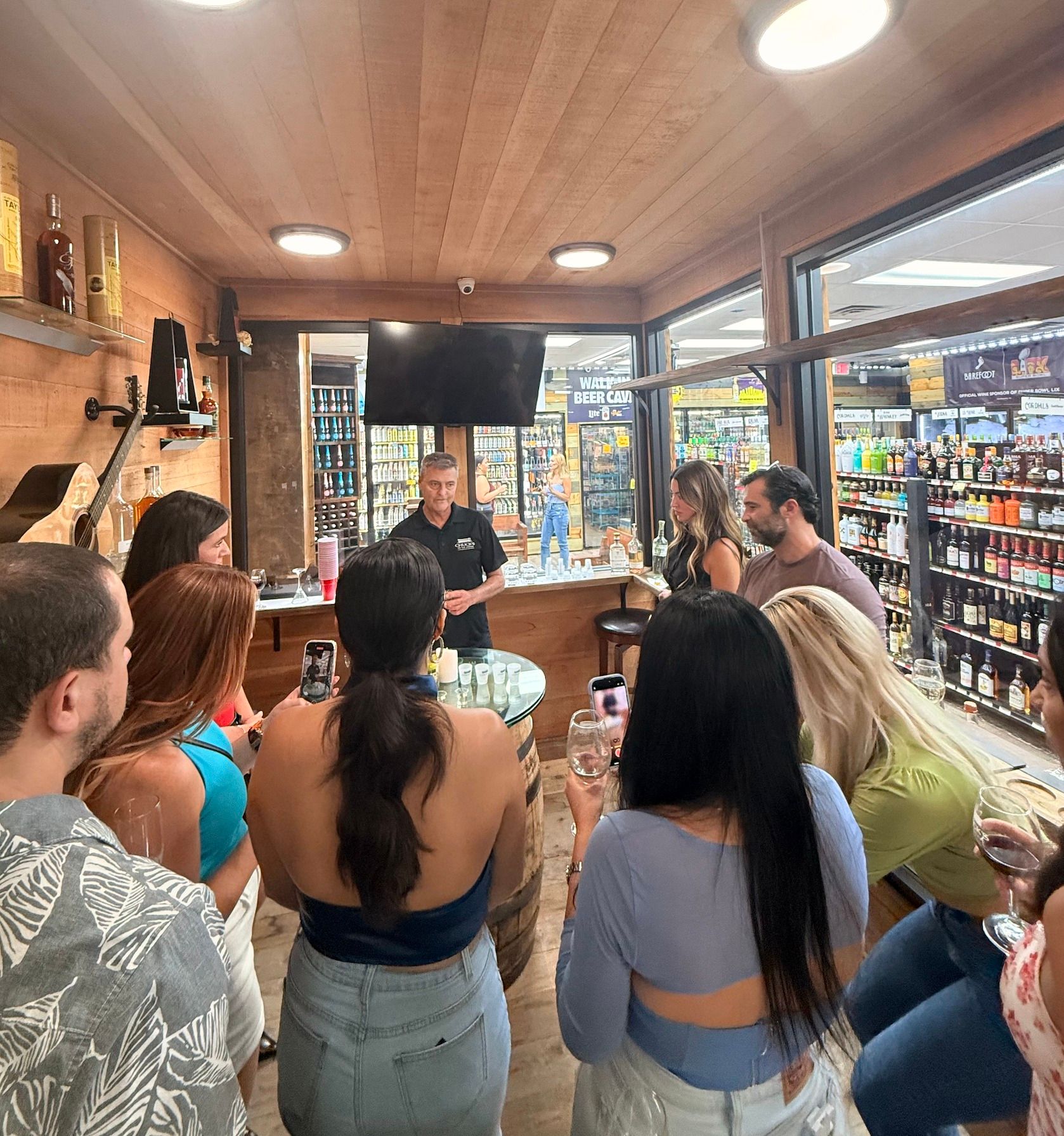 Crowd at a liquor store tasting room with warm wood interior, host pouring spirit samples as patrons hold wine glasses and phones near shelves of bottles visible through glass