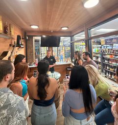 Crowd at a liquor store tasting room with warm wood interior, host pouring spirit samples as patrons hold wine glasses and phones near shelves of bottles visible through glass