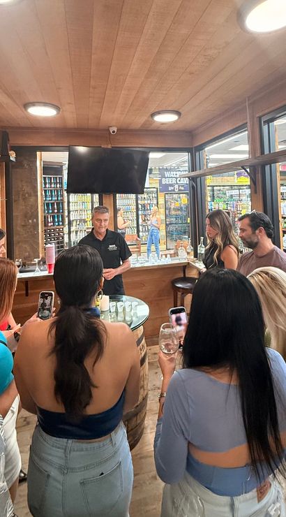 Crowd at a liquor store tasting room with warm wood interior, host pouring spirit samples as patrons hold wine glasses and phones near shelves of bottles visible through glass