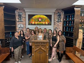 Bachelorette party group of women in leopard-print outfits posing inside a cozy wine shop tasting room with brick walls, arched stained-glass window, wooden wine racks and a carved lectern.