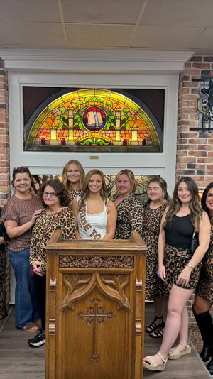 Bachelorette party group of women in leopard-print outfits posing inside a cozy wine shop tasting room with brick walls, arched stained-glass window, wooden wine racks and a carved lectern.