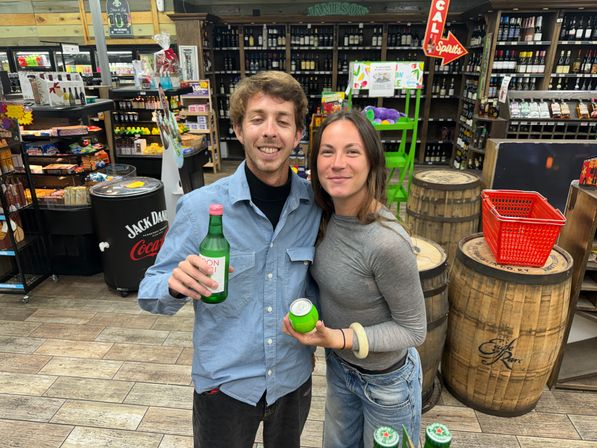 Two smiling adults posing inside a liquor store holding green bottled and canned drinks, standing by wooden barrels, wine shelves and a red shopping basket.