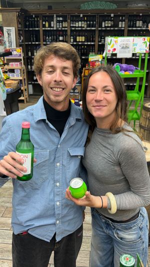 Two smiling adults posing inside a liquor store holding green bottled and canned drinks, standing by wooden barrels, wine shelves and a red shopping basket.
