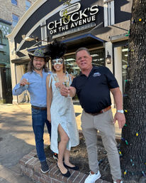 Three people toasting with champagne on a sunny downtown sidewalk outside a wine-and-spirits storefront — woman in a white dress and feathered masquerade mask, one man in a glittery cowboy hat, another in a black polo, tree wrapped in colorful string lights.