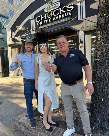 Three people toasting with champagne on a sunny downtown sidewalk outside a wine-and-spirits storefront — woman in a white dress and feathered masquerade mask, one man in a glittery cowboy hat, another in a black polo, tree wrapped in colorful string lights.