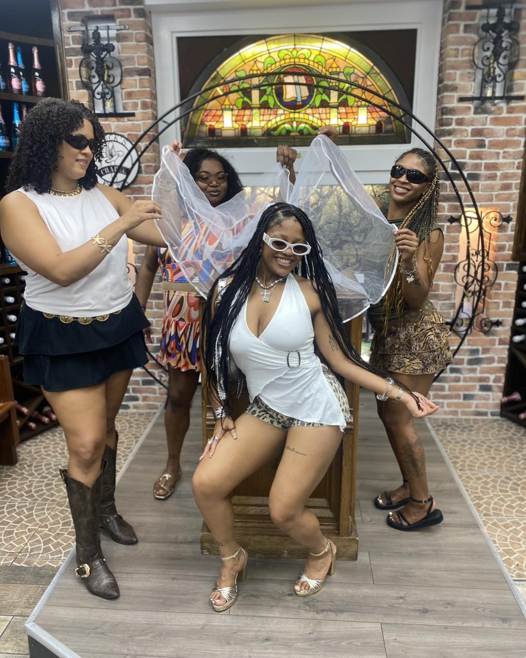 Four women enjoying a bachelorette celebration in a brick‑walled wine shop tasting room; bride‑to‑be seated in white with a sheer veil while friends pose, smile and wear sunglasses.