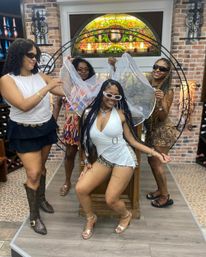 Four women enjoying a bachelorette celebration in a brick‑walled wine shop tasting room; bride‑to‑be seated in white with a sheer veil while friends pose, smile and wear sunglasses.
