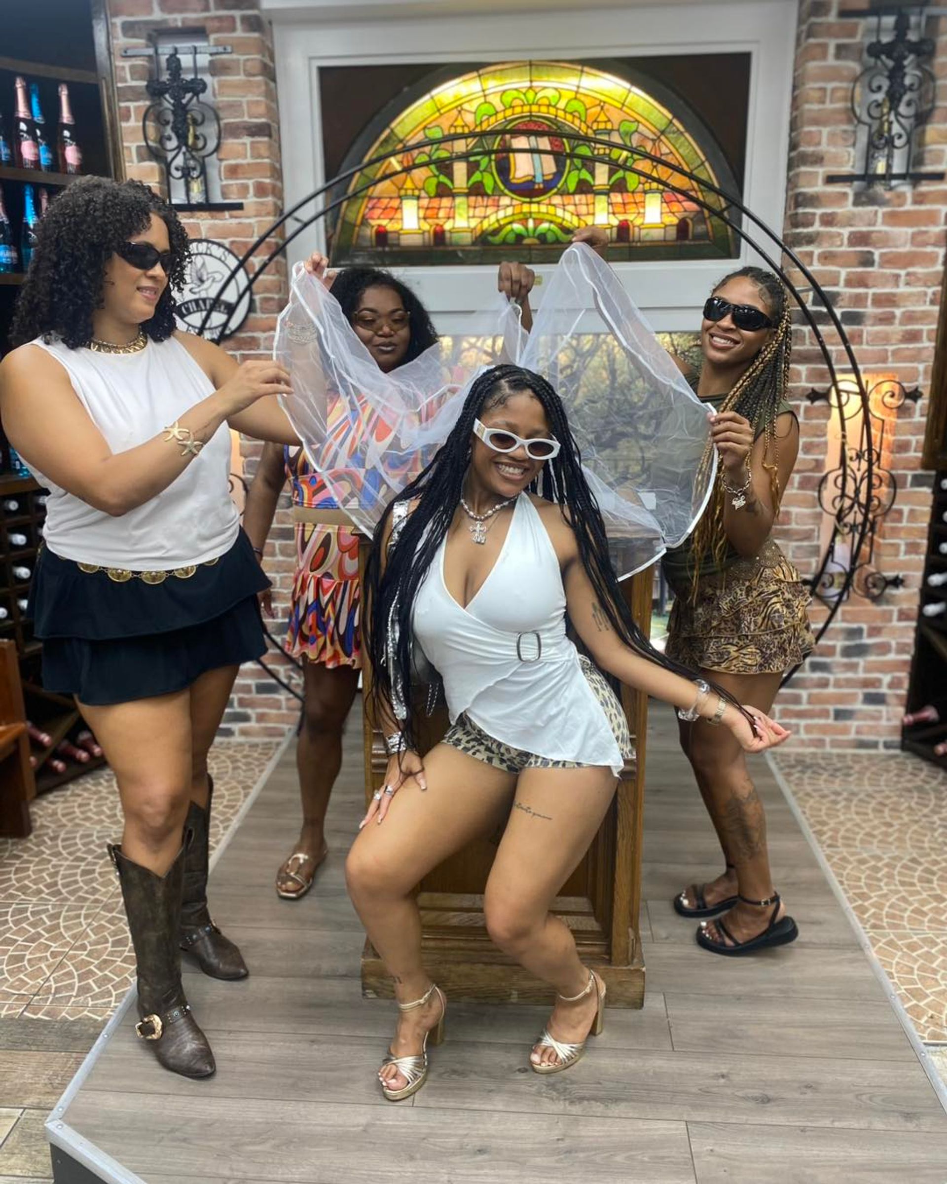 Four women enjoying a bachelorette celebration in a brick‑walled wine shop tasting room; bride‑to‑be seated in white with a sheer veil while friends pose, smile and wear sunglasses.