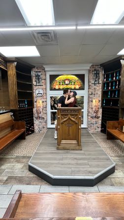 Couple kissing behind a carved wooden pulpit on a small raised indoor chapel display with stained-glass window, exposed brick walls, wooden pews and shelves holding bottles.