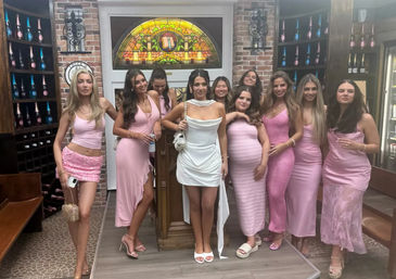Group photo of women in pink dresses and one in white posing on a small stage inside a cozy wine-bar style interior with exposed brick, arched stained-glass window and shelves of sparkling wine bottles — fun girls' night out