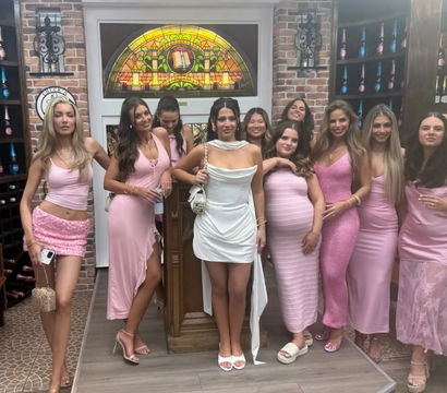 Group photo of women in pink dresses and one in white posing on a small stage inside a cozy wine-bar style interior with exposed brick, arched stained-glass window and shelves of sparkling wine bottles — fun girls' night out