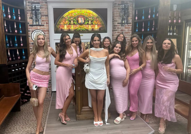 Group photo of women in pink dresses and one in white posing on a small stage inside a cozy wine-bar style interior with exposed brick, arched stained-glass window and shelves of sparkling wine bottles — fun girls' night out