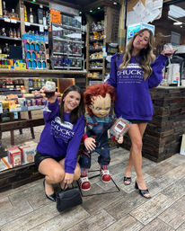 Two friends in matching purple sweatshirts toast with red wine while posing with a Chucky doll in a liquor store/wine shop aisle surrounded by bottles and snacks.