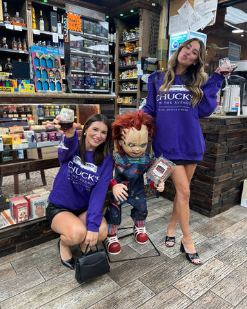 Two friends in matching purple sweatshirts toast with red wine while posing with a Chucky doll in a liquor store/wine shop aisle surrounded by bottles and snacks.