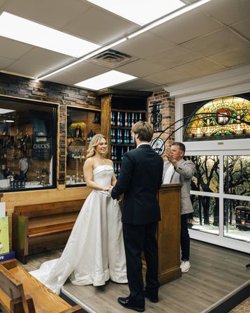 Smiling bride in a strapless white gown and groom in a black suit exchange vows at an intimate indoor micro-wedding in a cozy boutique shop with stained-glass window and wooden shelving.