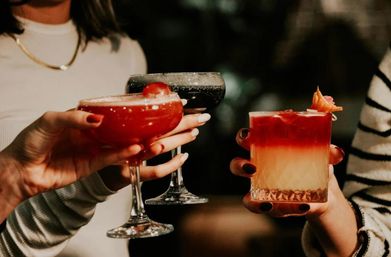 Three colorful cocktails raised in a toast at a dim bar: a red coupe, a dark coupe, and a layered orange-red rocks glass with a citrus garnish.