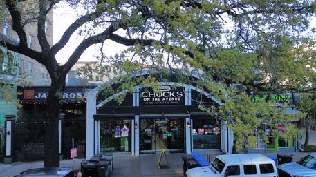 Tree-lined city avenue with a small wine and spirits storefront flanked by neighboring shops, neon window signs, parked cars and sidewalk bins.