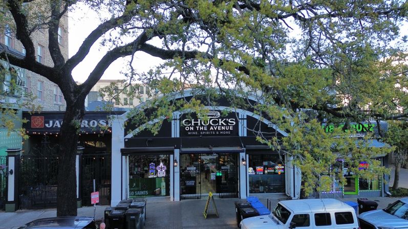 Tree-lined city avenue with a small wine and spirits storefront flanked by neighboring shops, neon window signs, parked cars and sidewalk bins.