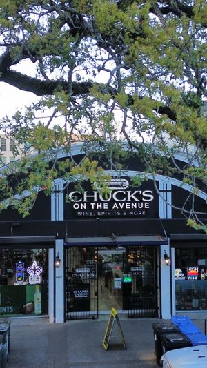 Tree-lined city avenue with a small wine and spirits storefront flanked by neighboring shops, neon window signs, parked cars and sidewalk bins.
