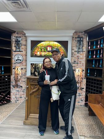 Smiling couple in matching black Adidas tracksuits pose inside a cozy wine shop-style tasting room with exposed brick walls, arched stained-glass window, wooden wine racks of colorful bottles, and a carved lectern; woman holding a takeout cup.