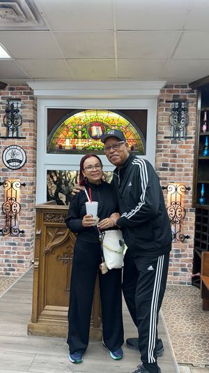 Smiling couple in matching black Adidas tracksuits pose inside a cozy wine shop-style tasting room with exposed brick walls, arched stained-glass window, wooden wine racks of colorful bottles, and a carved lectern; woman holding a takeout cup.