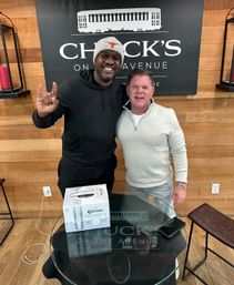 Two men smiling inside a wood-paneled interior, one in a beanie flashing a hand-horns sign; a glass table in front holds a 12-pack of Corona Premier beer and a smartphone.
