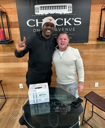 Two men smiling inside a wood-paneled interior, one in a beanie flashing a hand-horns sign; a glass table in front holds a 12-pack of Corona Premier beer and a smartphone.