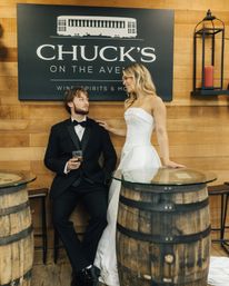 Bride in a strapless white wedding gown and groom in a black tuxedo pose at a rustic wood‑paneled tasting bar with whiskey barrels and a dark wall sign, wedding reception vibe.
