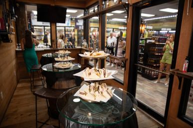 Bite-sized tea sandwiches on a three-tier stand atop glass tables in a rustic liquor-store tasting room, with shelves of wine and spirits and shoppers visible in the background.