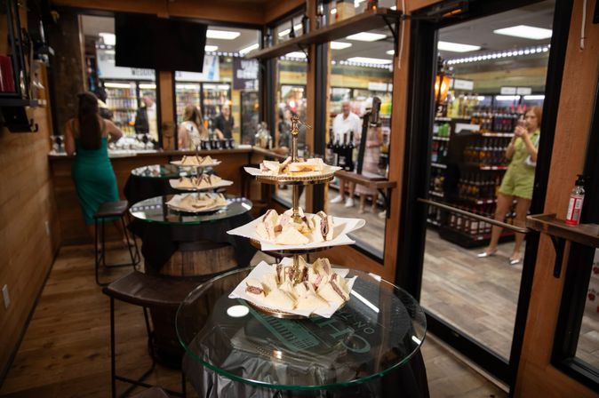 Bite-sized tea sandwiches on a three-tier stand atop glass tables in a rustic liquor-store tasting room, with shelves of wine and spirits and shoppers visible in the background.