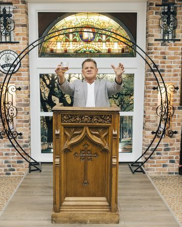Man raising a glass and gesturing from a carved wooden pulpit with a cross, stained-glass window and brick-walled chapel-style interior with decorative iron accents.